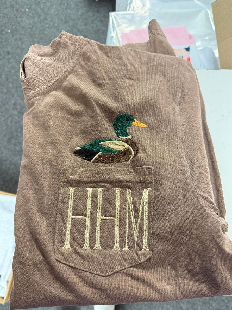 A folded brown t-shirt featuring an embroidered mallard duck on the chest and a front pocket with 'HHM' embroidered in beige.