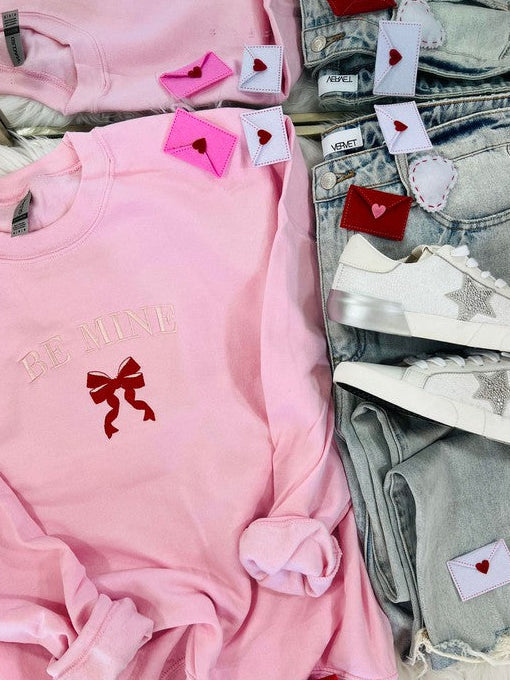 The image displays a light pink embroidered sweatshirt with the phrase "BE MINE" and a red ribbon design, presented next to a pair of blue denim jeans. The sweatshirt features decorative red heart-shaped tags and embroidered stars, enhancing its Valentine's Day-themed aesthetic.