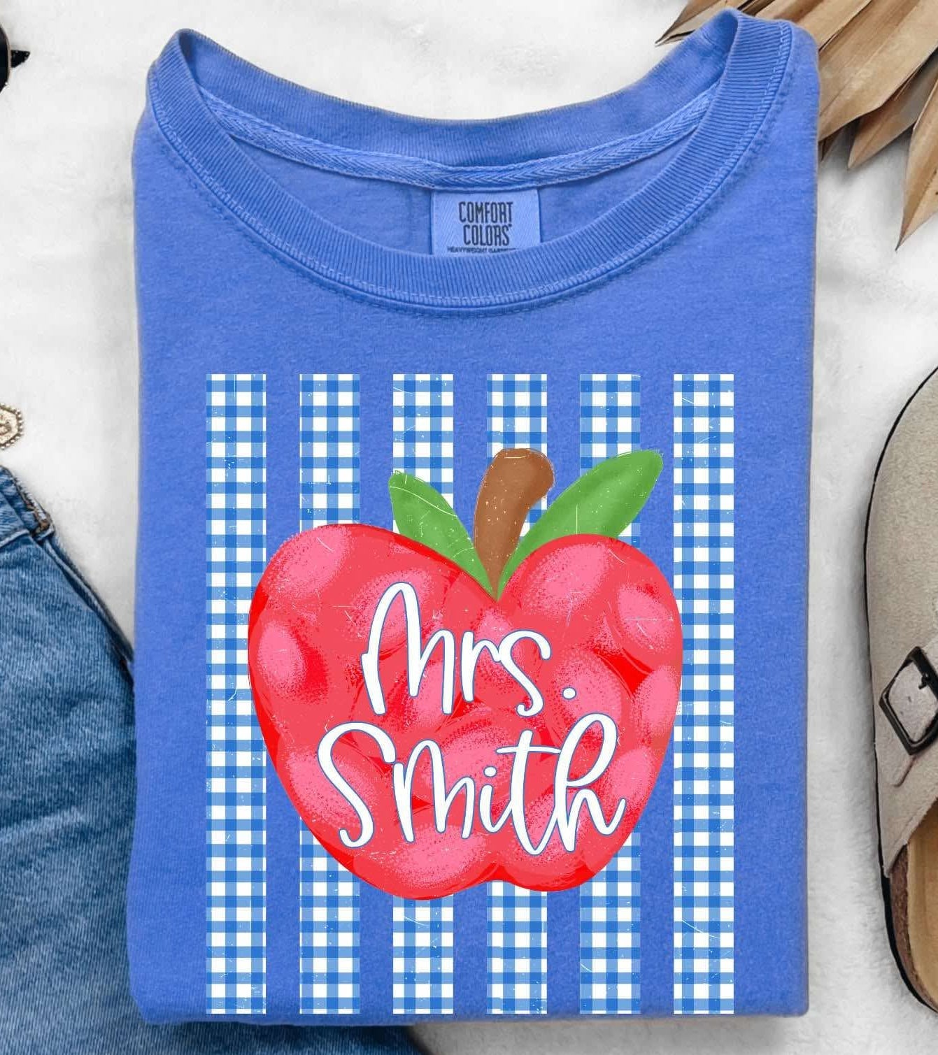 A folded blue T-shirt with a red apple graphic and the text 'Mrs. Smith' over blue-and-white gingham stripes, styled in a flat lay with jeans, sunglasses, a necklace, a slip-on shoe, and dried palm leaves on a white textured background.