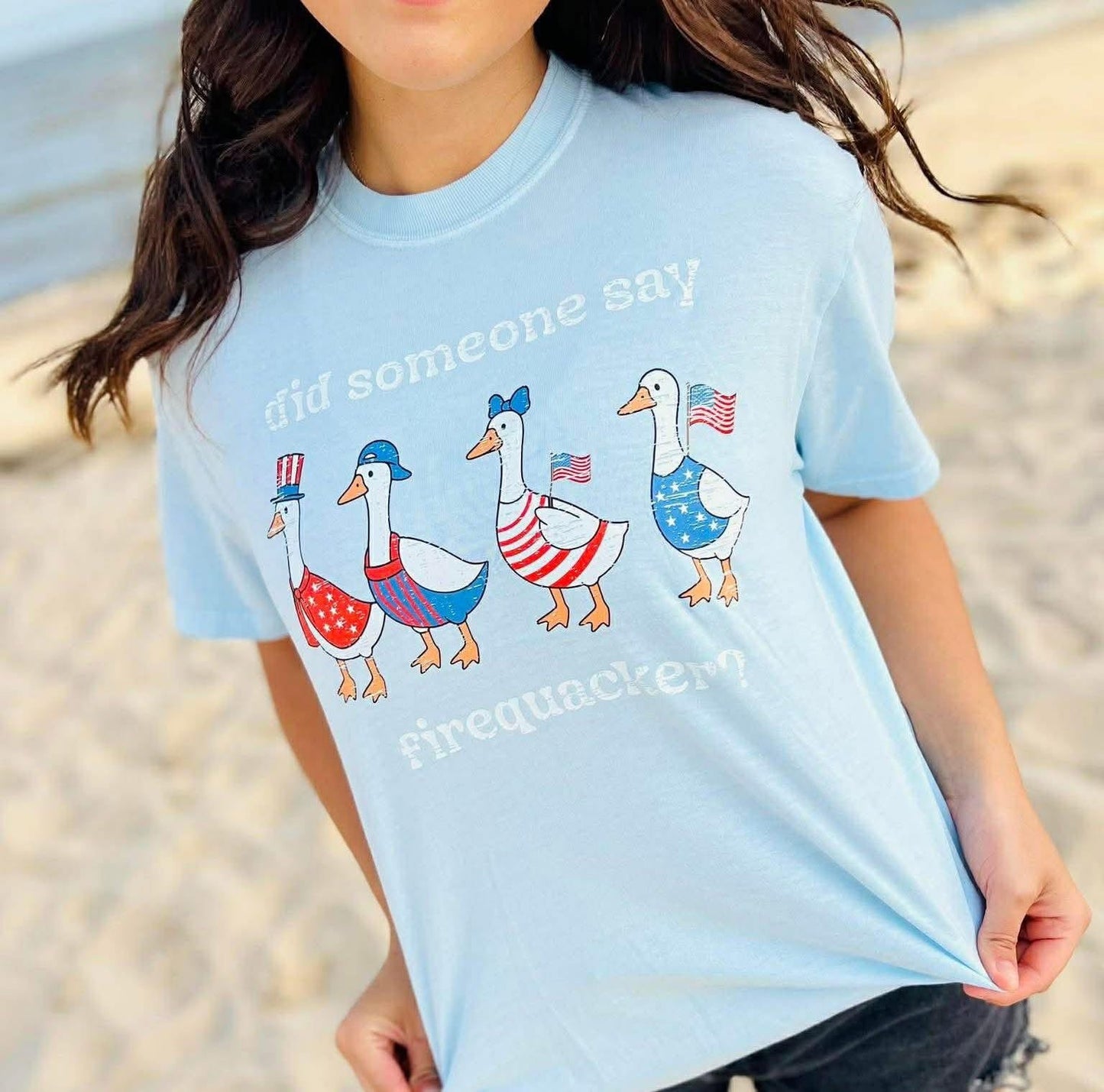 Light blue t-shirt with patriotic duck graphics worn by a person on a beach.